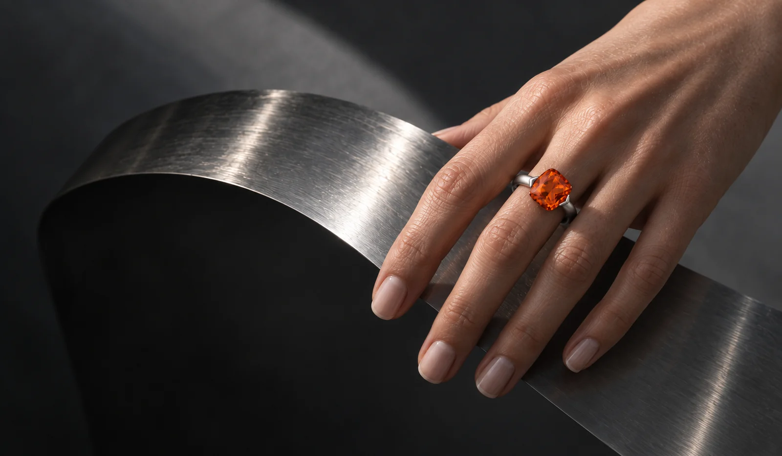 An orange gemstone ring worn against a curved, brushed-steel sculpture
